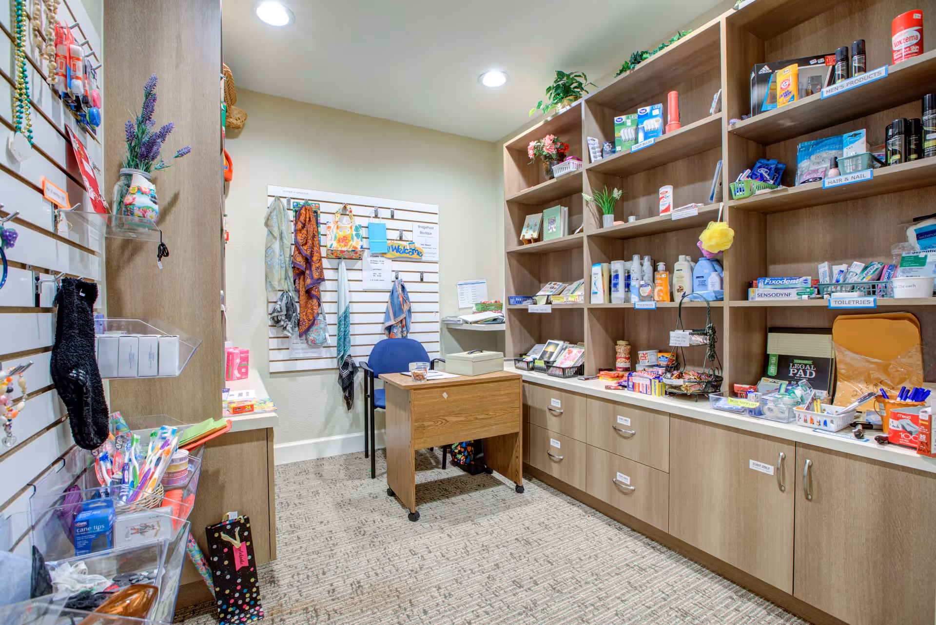 A small retail or convenience shop area inside a senior living facility with wooden shelves and cabinets stocked with personal care items, toiletries, snacks, and small accessories. A wooden desk with a blue chair is positioned in the center, and various scarves and items hang on a white slat wall. The room is well-lit with recessed ceiling lights and has a carpeted floor.