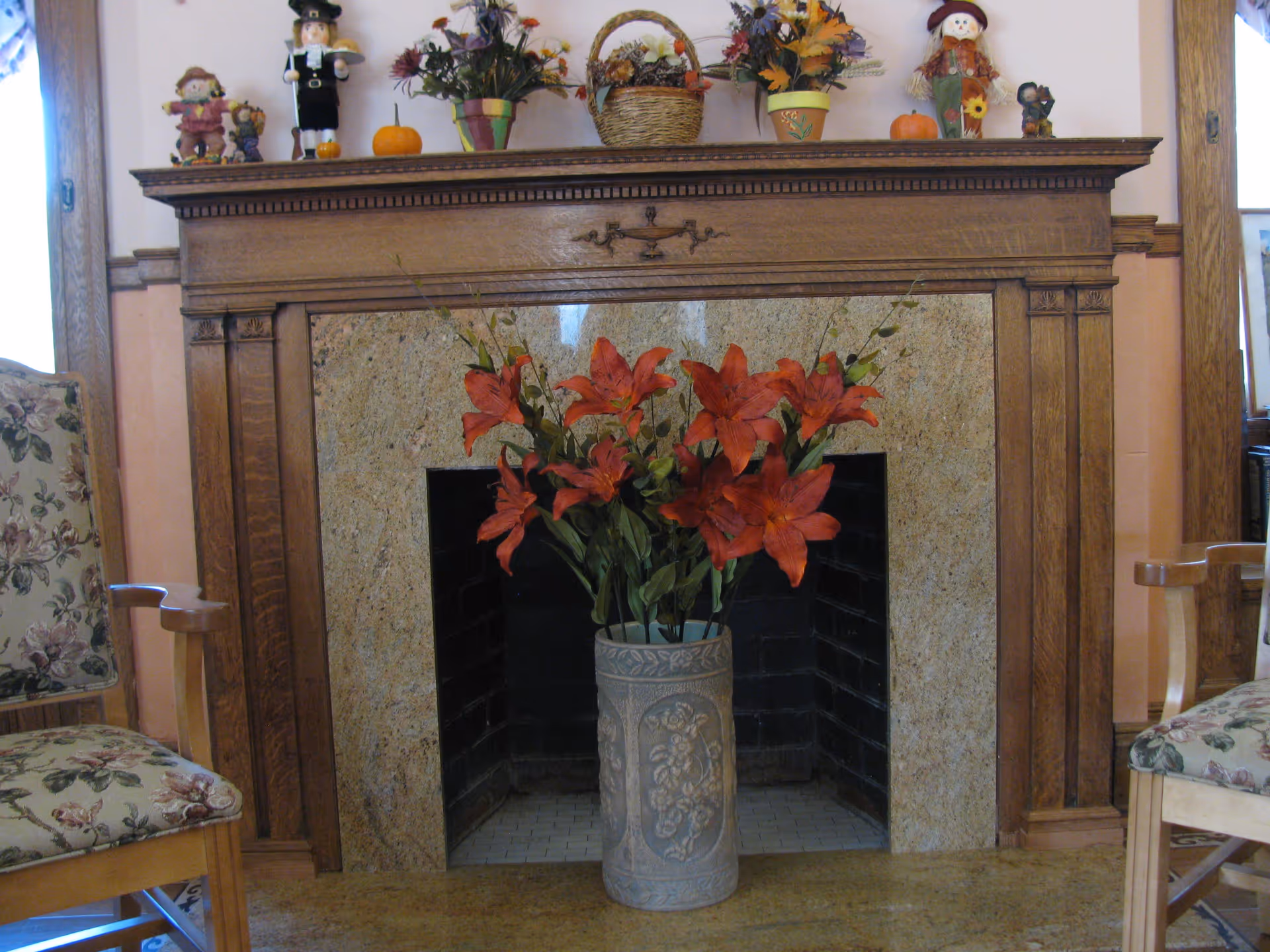 A decorative fireplace with a wooden mantel adorned with small autumn-themed figurines, pumpkins, and potted plants. In front of the fireplace is a large vase filled with vibrant red flowers. Two floral upholstered chairs are partially visible on either side of the fireplace.