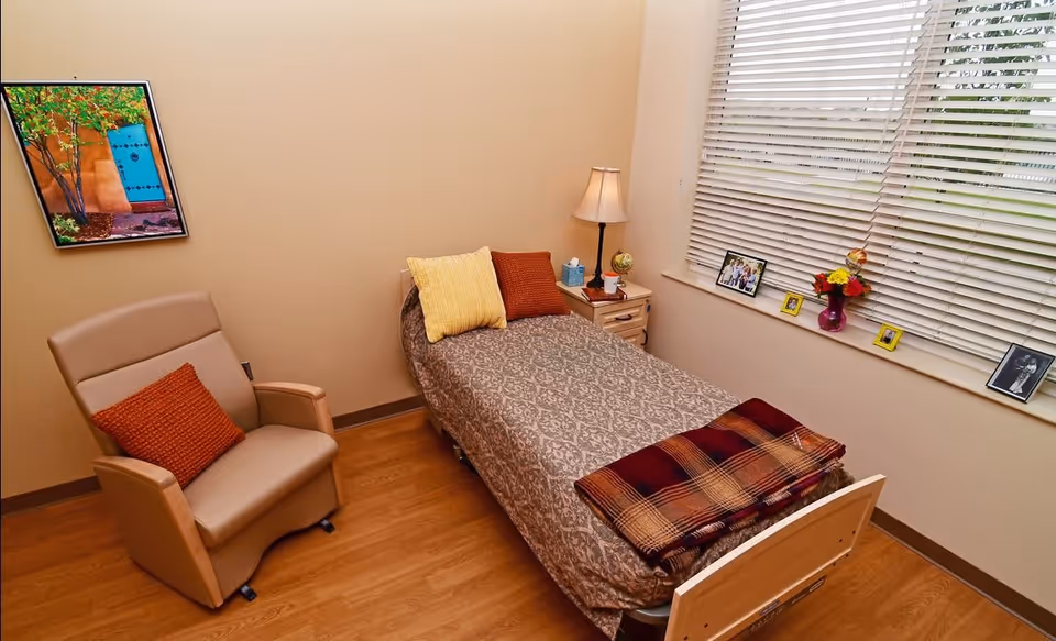 A small, cozy bedroom with a single bed covered in a patterned bedspread and two pillows, one yellow and one orange. A plaid blanket is folded at the foot of the bed. Next to the bed is a wooden nightstand with a lamp, a tissue box, a small globe, and a cup. A comfortable beige armchair with an orange pillow is positioned near the bed. The room has wooden flooring, a large window with white blinds, and several framed photos and a vase with flowers on the windowsill. A colorful framed picture hangs on the wall.
