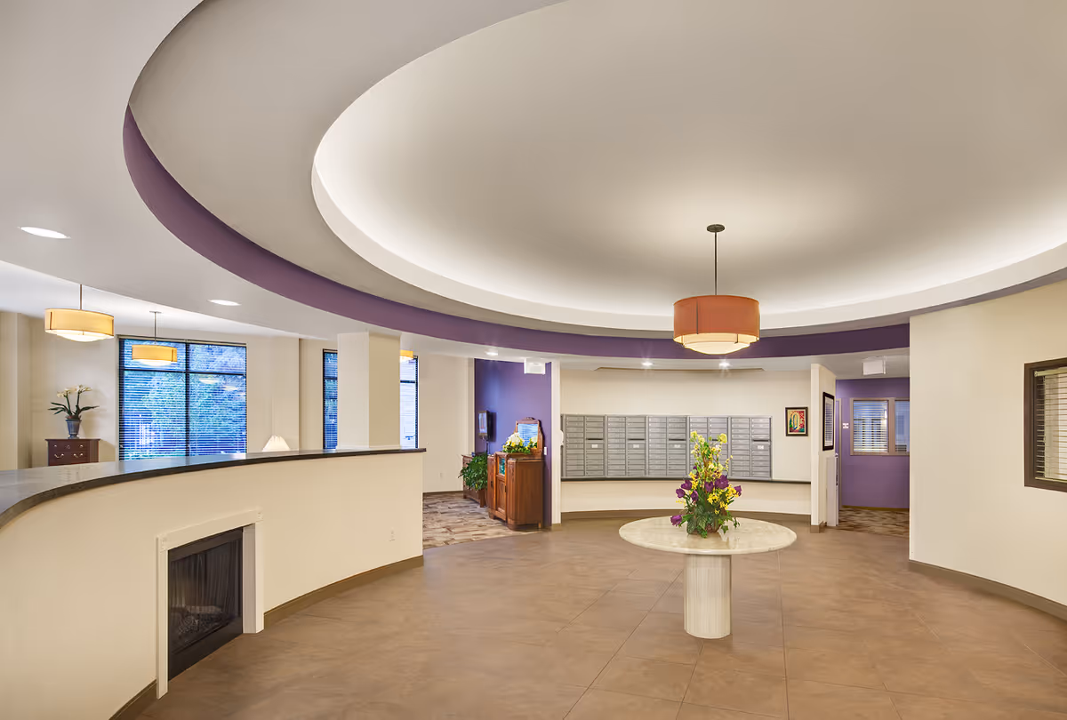 Interior view of a senior living facility lobby area with a circular ceiling design featuring recessed lighting and a central hanging light fixture. A round table with a floral arrangement is in the center of the tiled floor. Mailboxes are mounted on the far wall, and there are windows and doorways leading to other rooms. The color scheme includes beige walls with purple accents.