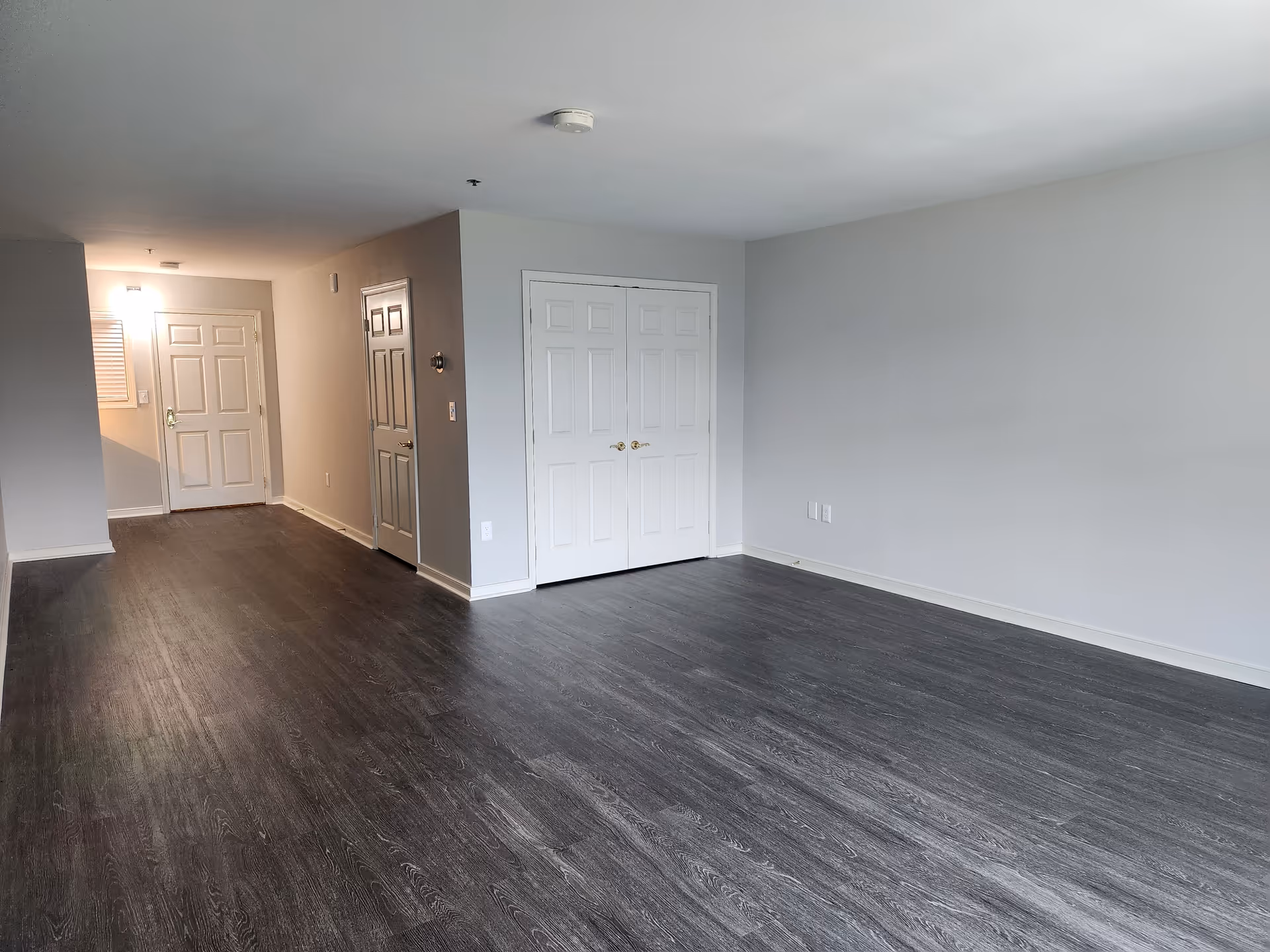 Empty room with dark wood flooring, light gray walls, and white doors including a closet and an entrance door. The room is well-lit with ceiling lights and has a simple, clean appearance.