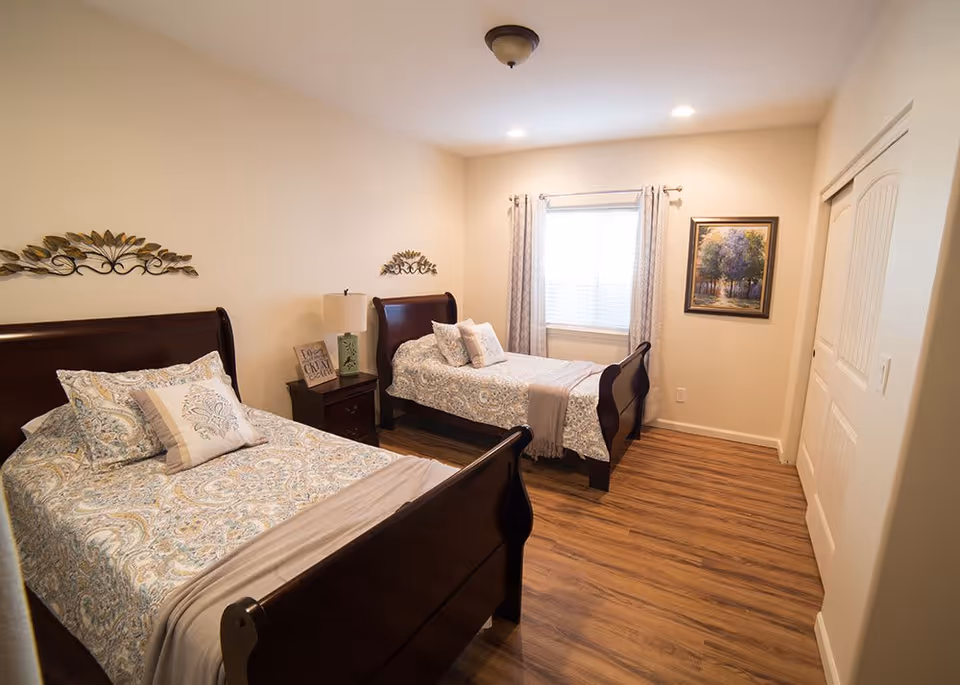 A bedroom with two twin beds featuring dark wooden frames and patterned bedding. Between the beds is a nightstand with a lamp and decorative items. The room has light-colored walls, a window with curtains, a framed painting on the wall, and wood flooring.