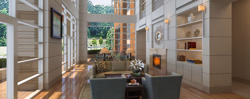 A spacious and well-lit living room with large floor-to-ceiling windows allowing natural light to fill the space. The room features a fireplace with a decorative mirror above it, built-in shelves with books and decorative items, a sofa with patterned cushions, two armchairs, a coffee table with a flower arrangement, and two table lamps. Outside the windows, greenery and a brick wall are visible.