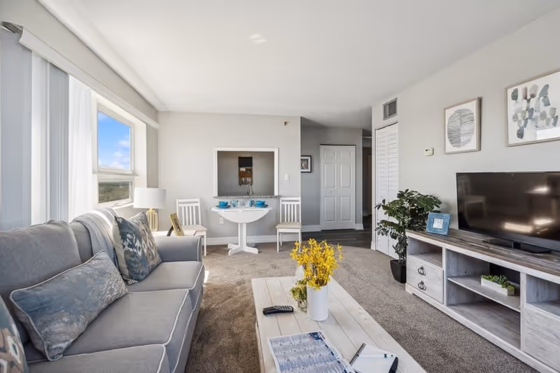A bright living room with a gray sofa adorned with patterned cushions, a wooden coffee table with a vase of yellow flowers, a TV on a wooden stand, two white chairs with a small round table set with blue dishes, and framed artwork on the walls. A large window lets in natural light.