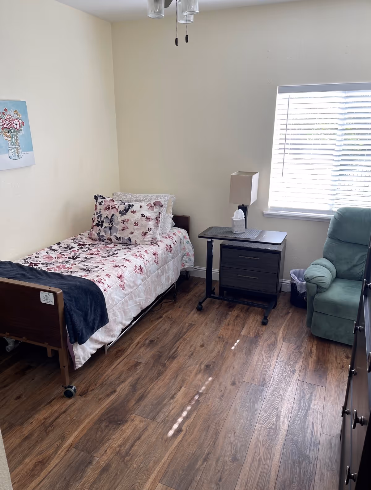 A small bedroom with a single bed covered in floral bedding, a bedside table with a lamp and tissue box, a green armchair, a window with blinds, and wooden flooring.