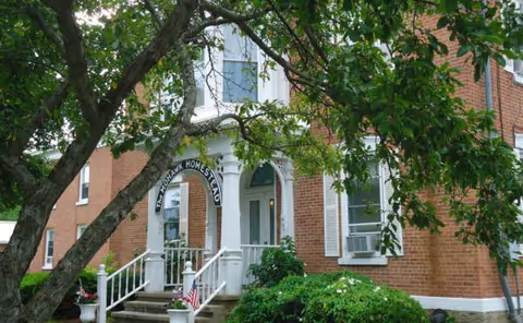 Brick senior living facility entrance with white trim, arched doorway, and surrounding greenery.