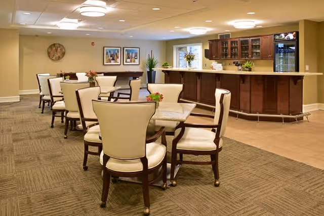 A well-lit common area with several tables and cream-colored cushioned chairs arranged around them. In the background, there is a wooden bar counter with glass cabinets and a small refrigerator. The walls are decorated with framed artwork and a round decorative piece. The floor is carpeted with a patterned design.