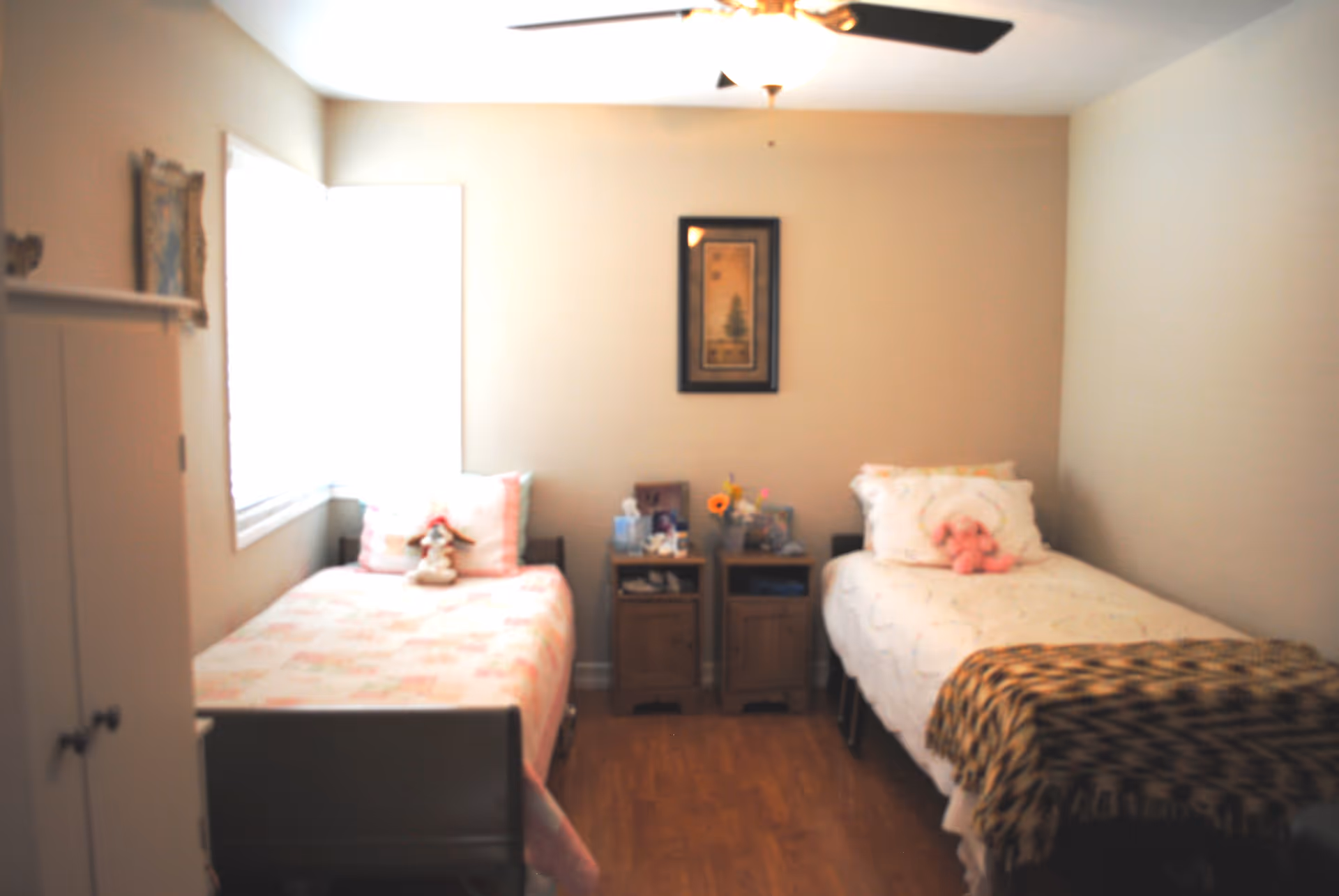 A small bedroom with two single beds placed parallel to each other against opposite walls. Each bed has pillows and stuffed animals on it. Between the beds are two wooden nightstands with various personal items and a framed picture hanging on the wall above them. The room has light-colored walls, a ceiling fan, and a window letting in natural light.