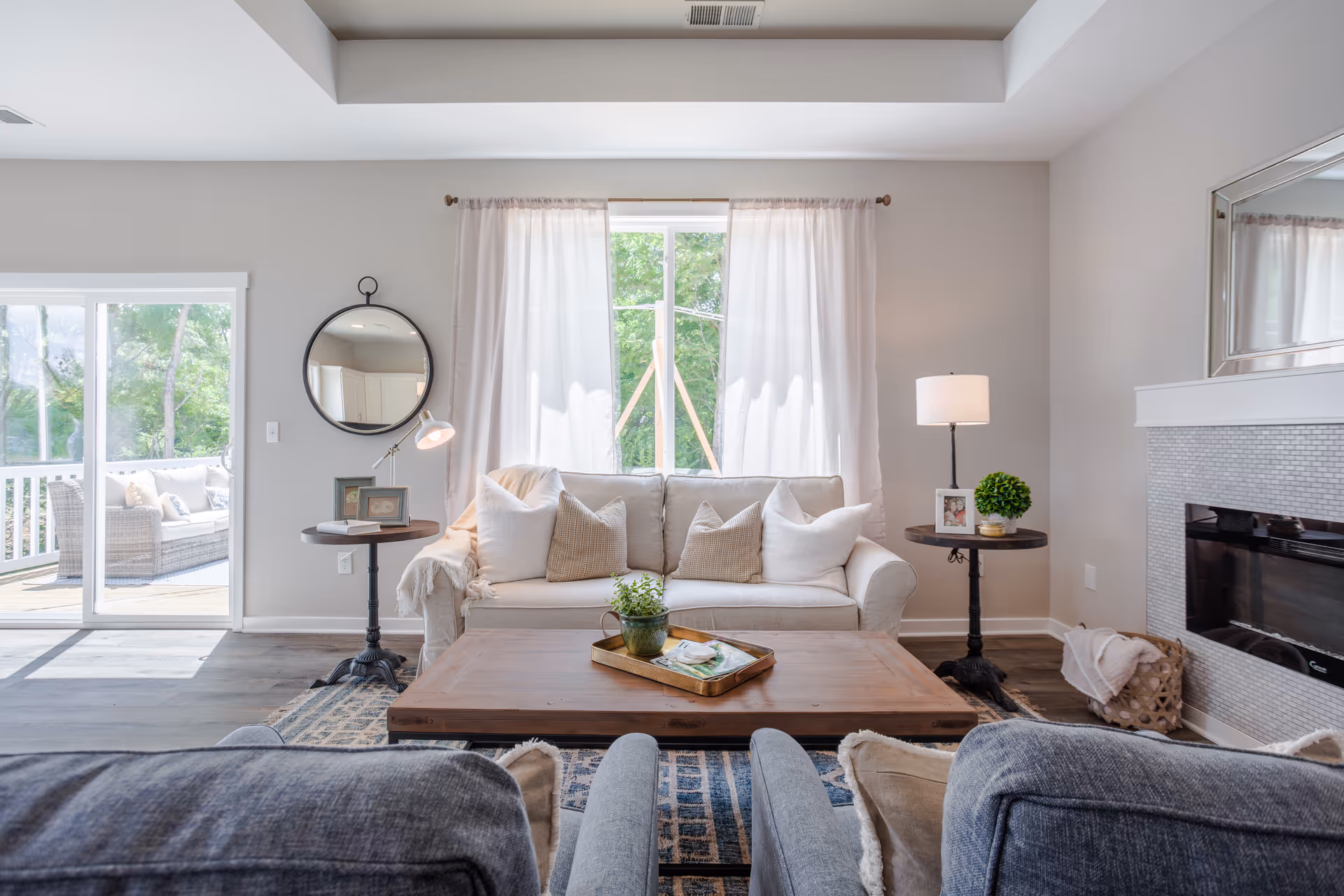 A bright and cozy living room with a white sofa adorned with beige and white pillows, a wooden coffee table with a small plant and tray, two side tables with lamps and framed photos, a round mirror on the wall, a large window with sheer white curtains, a fireplace with a basket of blankets, and sliding glass doors leading to an outdoor seating area.
