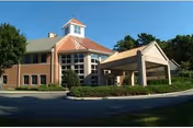 Front exterior of a brick senior living building with a covered entrance/porte-cochère, cupola, large windows, and landscaped driveway under a clear blue sky.