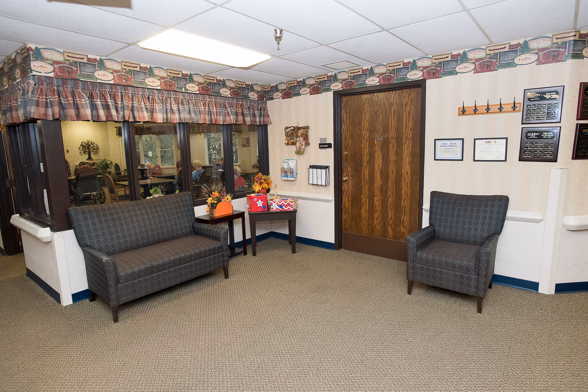 A waiting area in a senior living facility with a patterned loveseat and armchair, a small table with decorative items, a wooden door labeled 603, and a window showing people sitting inside another room. The walls have framed certificates and plaques, and the ceiling has white tiles.
