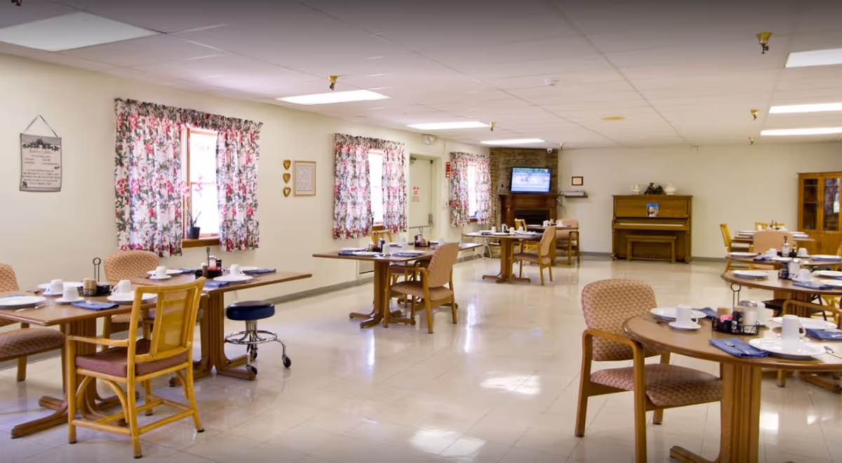 Bright communal dining room with multiple set tables and chairs, floral curtains, a piano and a TV at the far wall.