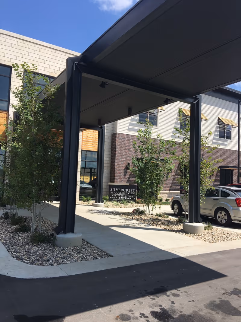 Entrance area of Silvercrest at College View Senior Living facility showing a covered drop-off zone with black metal supports, small trees planted in landscaped beds with rocks, and a sign indicating the main entrance. The building exterior features a mix of brick and light-colored siding with windows and yellow awnings. A silver car is parked nearby under a clear blue sky.