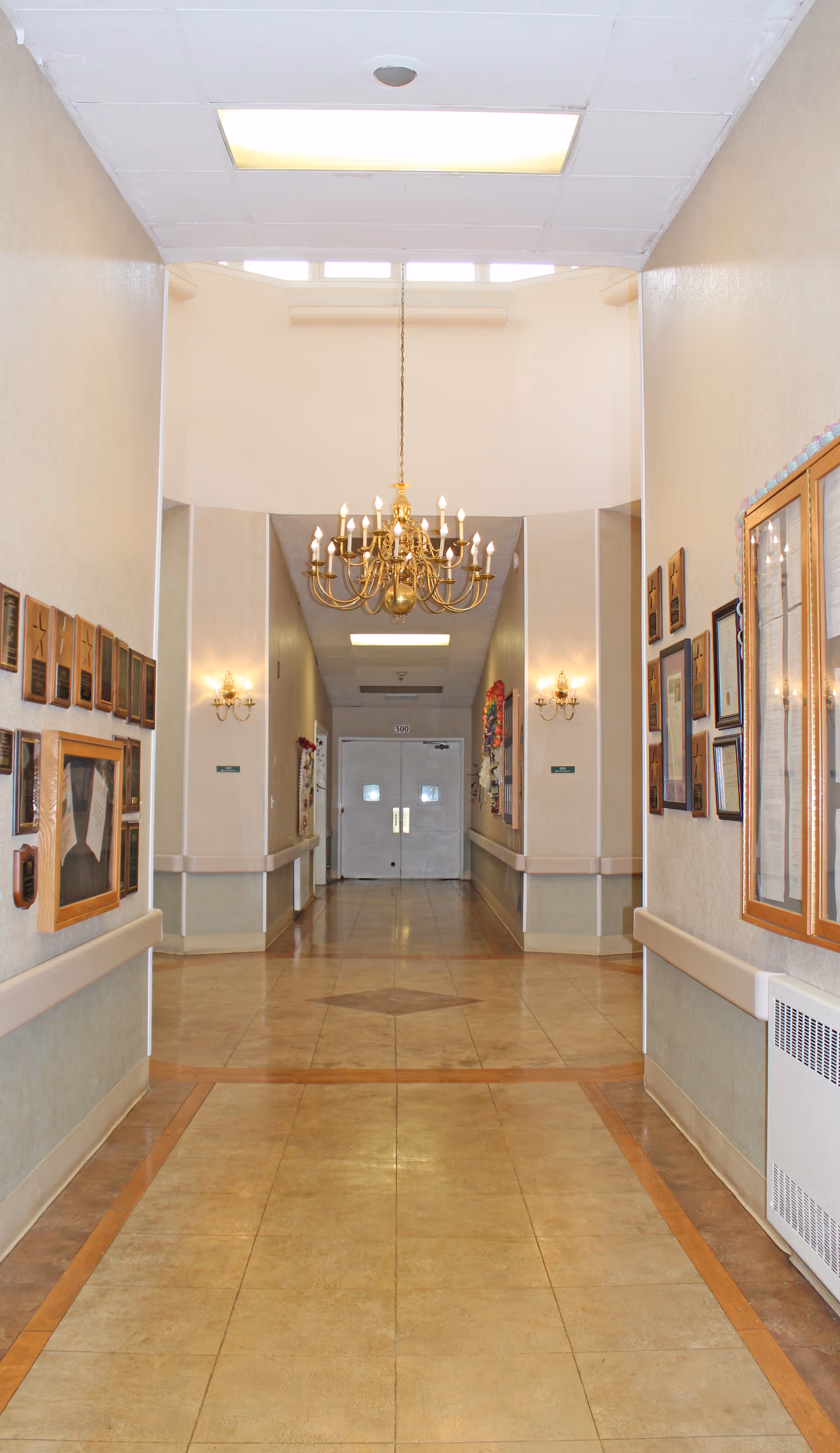 A well-lit hallway in a care and rehab center with beige tiled floors and cream-colored walls. The hallway is decorated with framed pictures and plaques on both sides. A large, ornate chandelier hangs from the ceiling, and there are wall sconces providing additional lighting. Double doors are visible at the end of the hallway.