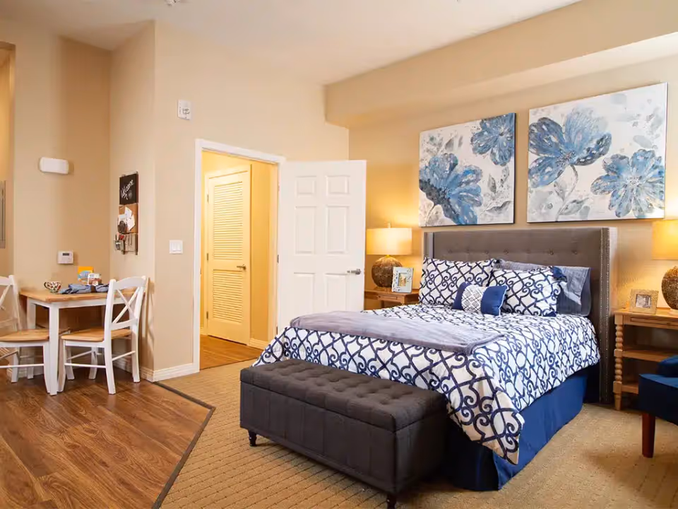 A cozy bedroom with a bed featuring blue and white patterned bedding, two large floral paintings above the headboard, two bedside tables with lamps, and a small dining table with chairs on the left side. The room has beige walls and a mix of carpet and wood flooring.