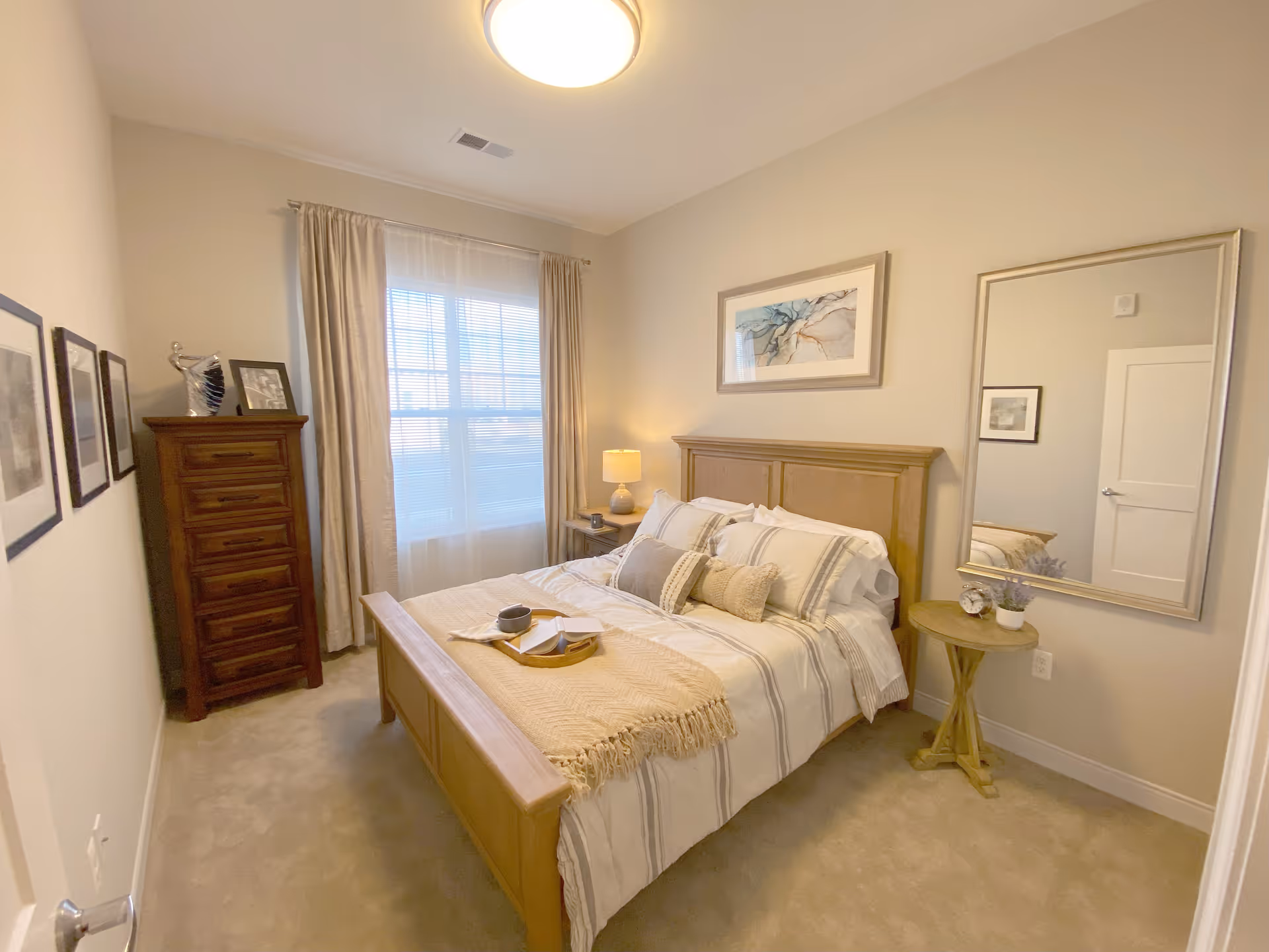 A cozy bedroom with a wooden bed frame and a neatly made bed with striped bedding and decorative pillows. A small wooden side table with a potted plant and a large mirror hangs on the wall next to the bed. On the opposite side, there is a tall wooden dresser with framed pictures and decorative items on top. A window with sheer curtains allows natural light to fill the room, and a ceiling light fixture illuminates the space.