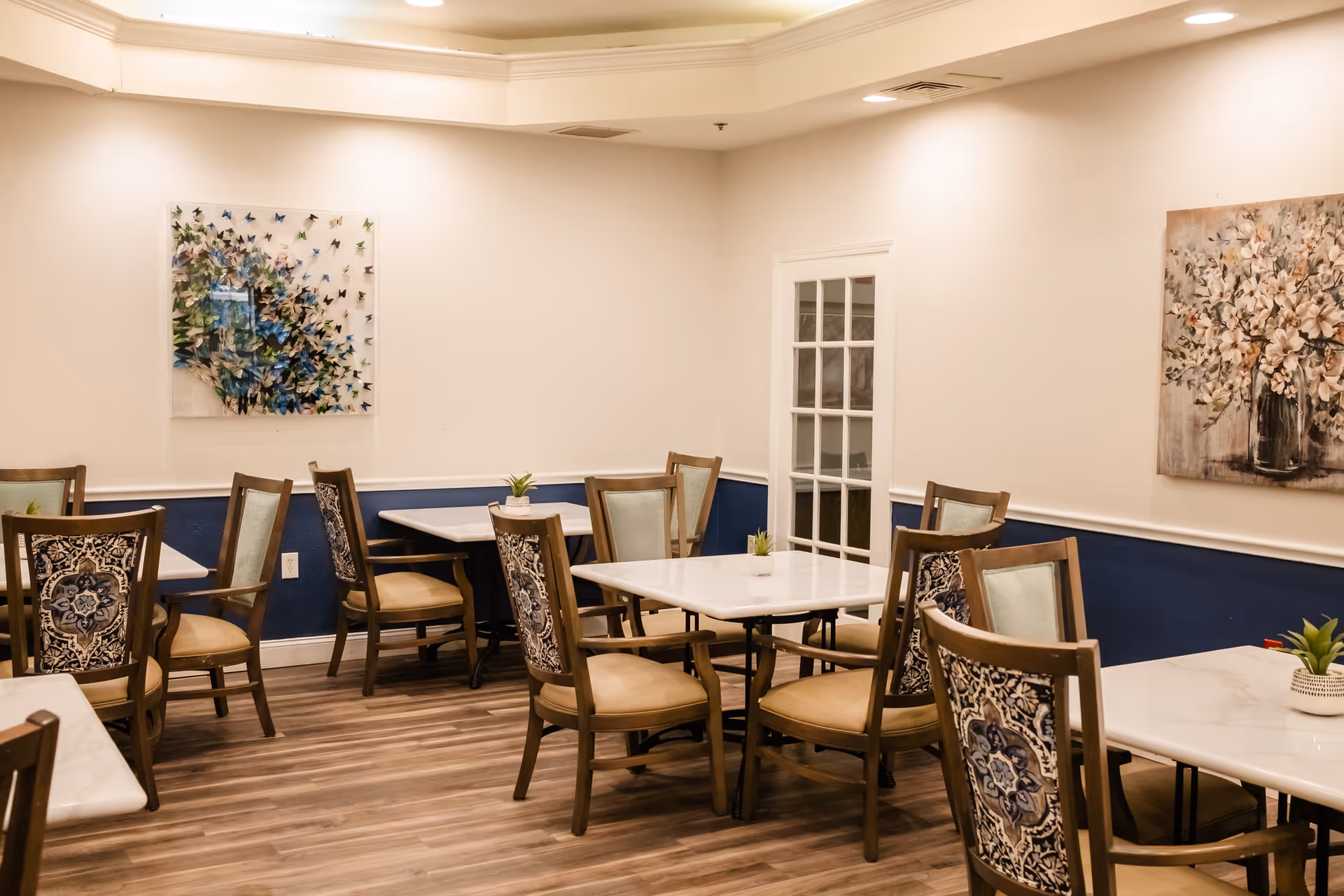 A dining room with several tables and chairs arranged neatly. The chairs have patterned backs and beige seats. The walls are painted white with a blue lower section, and there are two pieces of artwork hanging on the walls. The floor is wooden, and small potted plants are placed on the tables.