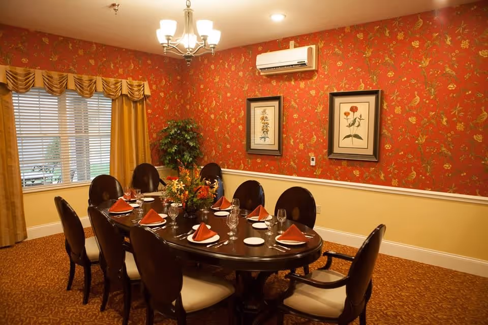 A formal dining room with a dark wooden oval table set for eight people. Each place setting has a white plate, a glass, and a folded orange napkin. The room features red floral wallpaper on the upper half of the walls and a yellow lower half with white trim. There are two framed botanical prints on the wall, a chandelier overhead, a window with yellow curtains, and a green potted plant in the corner.