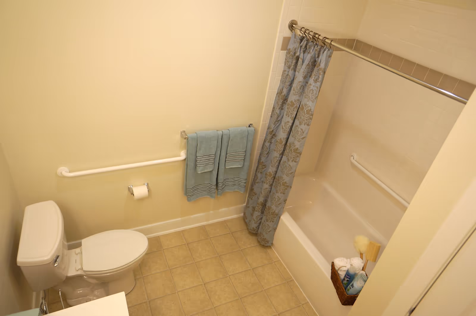 A clean bathroom with a white toilet, beige tiled floor, and a bathtub with a blue and beige patterned shower curtain. There are two blue towels hanging on a towel rack above a white grab bar, and a basket with bath supplies placed on the edge of the bathtub.