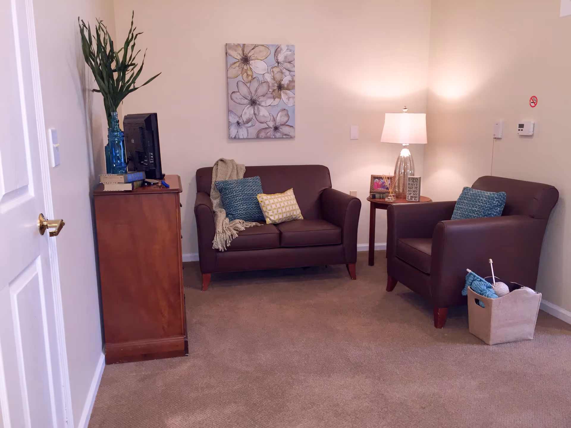 A cozy living room with a brown loveseat and matching armchair, each adorned with decorative pillows. A wooden side table with a lamp, framed photo, and decorative items sits between the chairs. A wooden cabinet with a TV and a tall vase with green plants is on the left side. A floral painting hangs on the wall above the loveseat. The room has beige walls and carpeted floor.