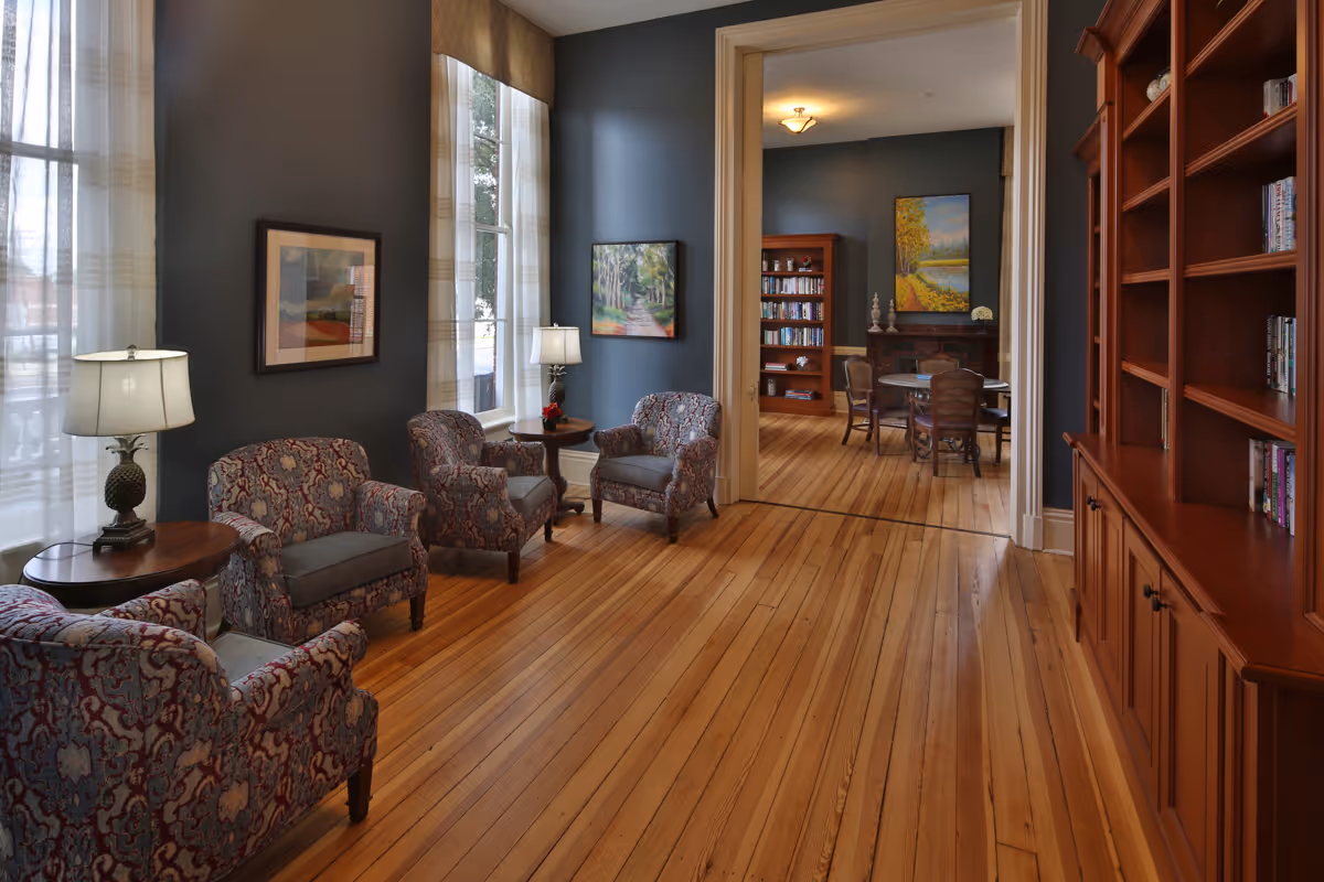 A cozy senior living common area with four patterned armchairs arranged around two small wooden side tables with lamps. The room has large windows with sheer curtains, blue-gray walls adorned with framed paintings, and polished wooden floors. In the background, an adjoining room features a round table with chairs, bookshelves, and a fireplace with a colorful landscape painting above it.