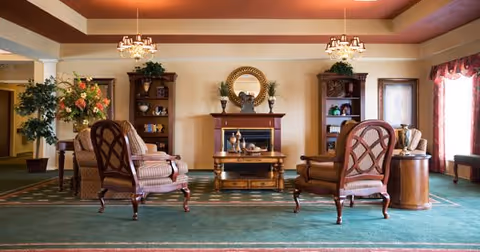 A cozy living room area with two wooden armchairs with patterned cushions facing a wooden coffee table and an electric fireplace. The fireplace has decorative items on the mantel and a round mirror above it. There are two wooden bookshelves on either side of the fireplace with plants and decorative items. The room has green carpet, beige walls, and two chandeliers hanging from the ceiling. Large windows with red curtains allow natural light into the room.