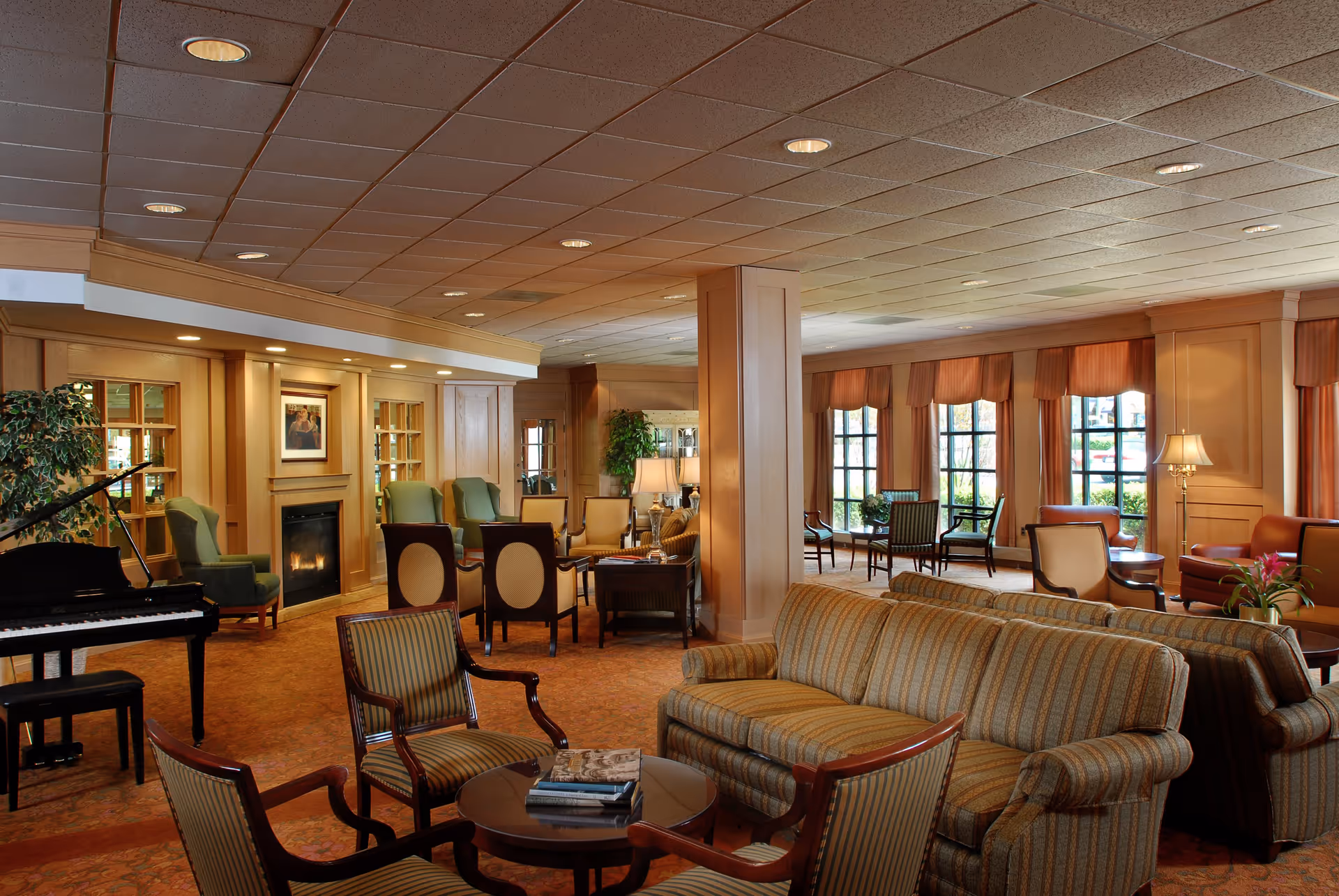 A spacious and warmly lit living room area in a senior living facility featuring multiple seating arrangements with sofas and armchairs, a grand piano on the left, a fireplace with a framed picture above it, large windows with curtains allowing natural light, and decorative plants and lamps throughout the room.