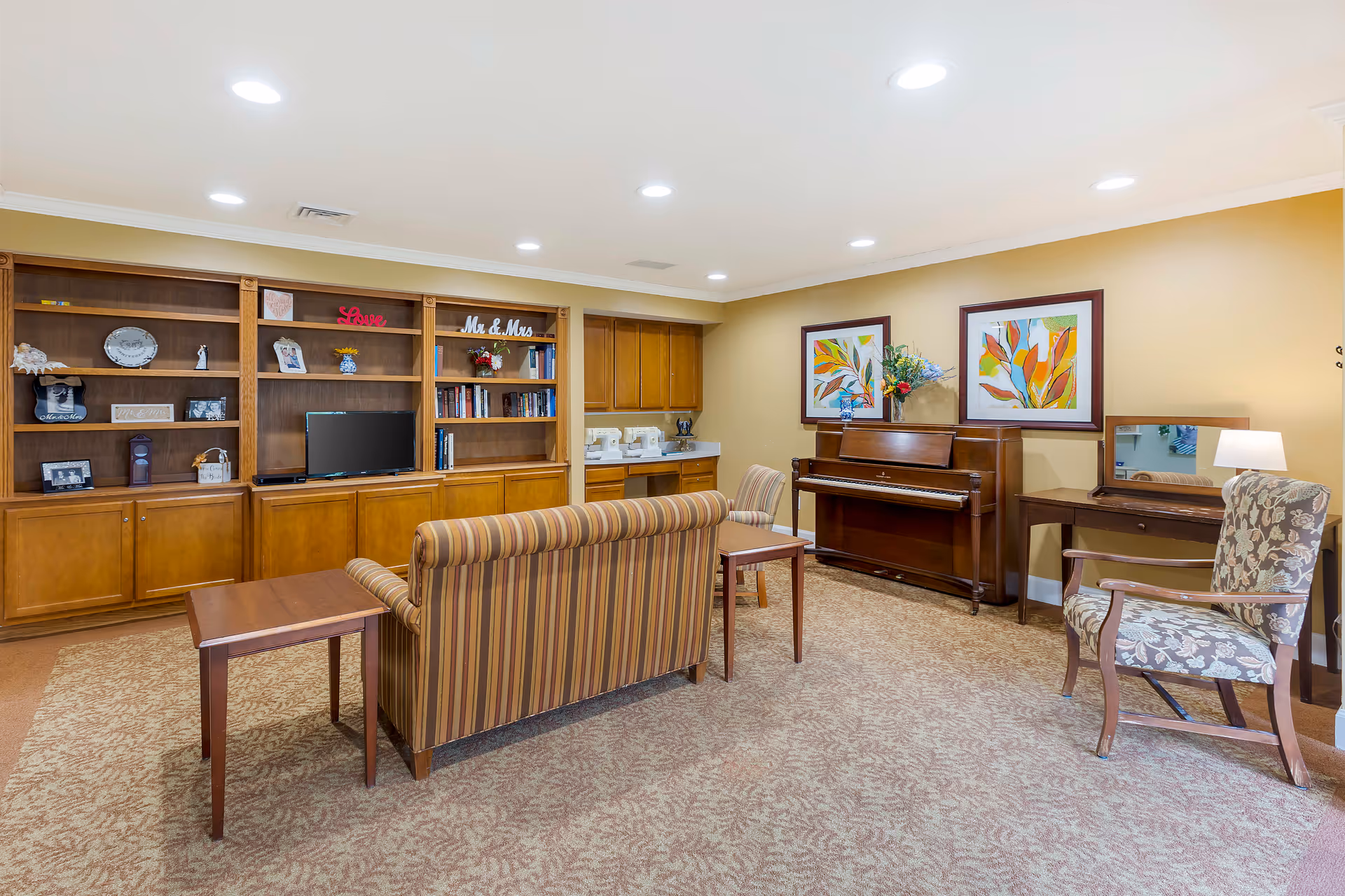 A cozy living room area with a striped sofa, wooden coffee table, floral armchair, and a wooden piano against a beige wall. There are two colorful framed paintings above the piano, a wooden desk with a mirror and lamp, and built-in wooden shelves with books, decorations, and a small TV. The room has recessed ceiling lights and carpeted flooring.