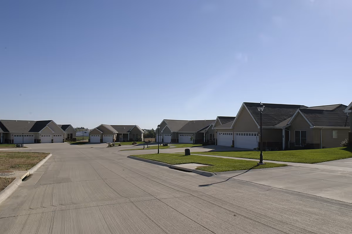 A quiet residential street in a senior living community with single-story houses featuring garages, green lawns, and street lamps under a clear blue sky.