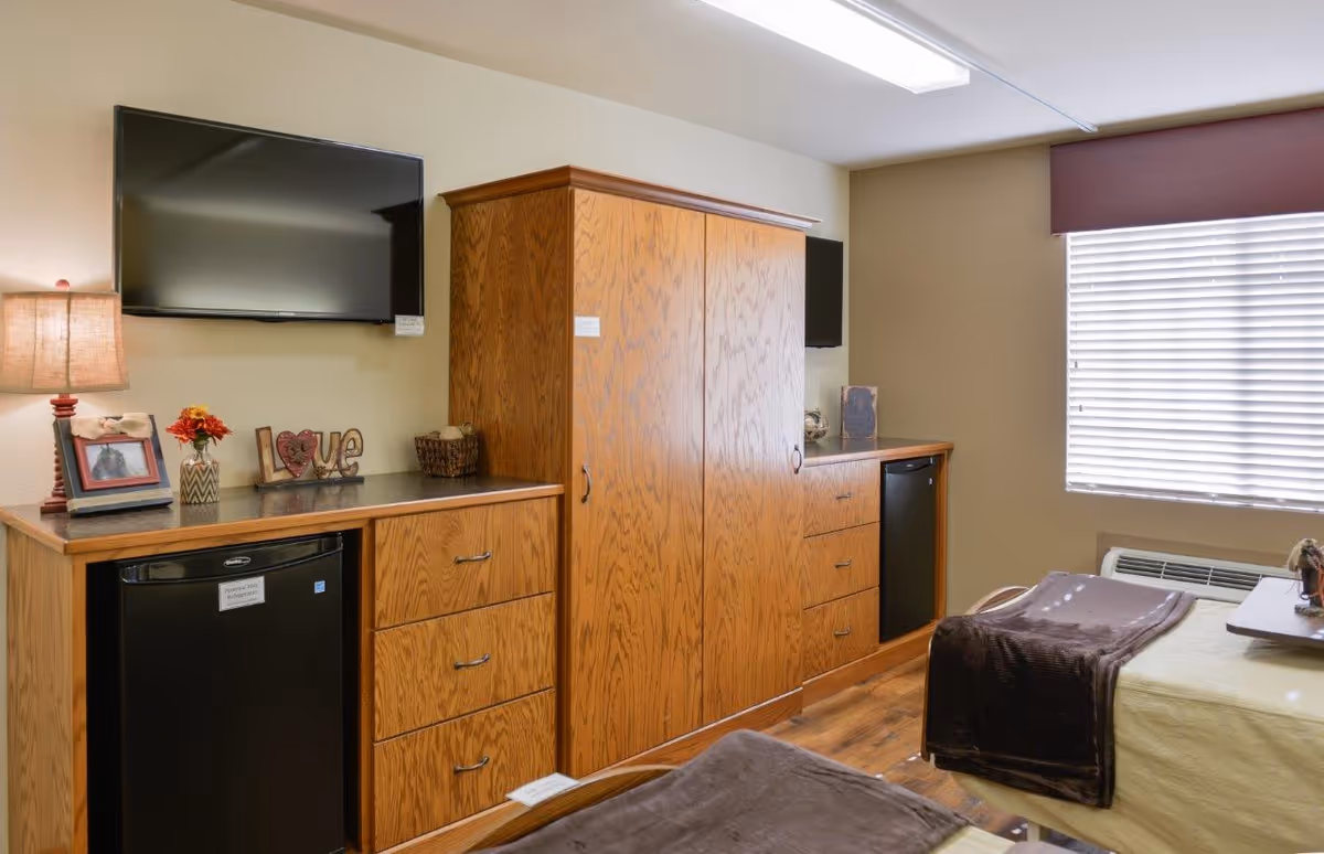 Interior view of a senior living facility room with two beds covered in beige and brown blankets. The room features wooden cabinets with drawers, two small refrigerators, a flat-screen TV mounted on the wall, a lamp, a small flower vase, and a decorative 'Love' sign on the cabinet. A window with blinds allows natural light into the room.