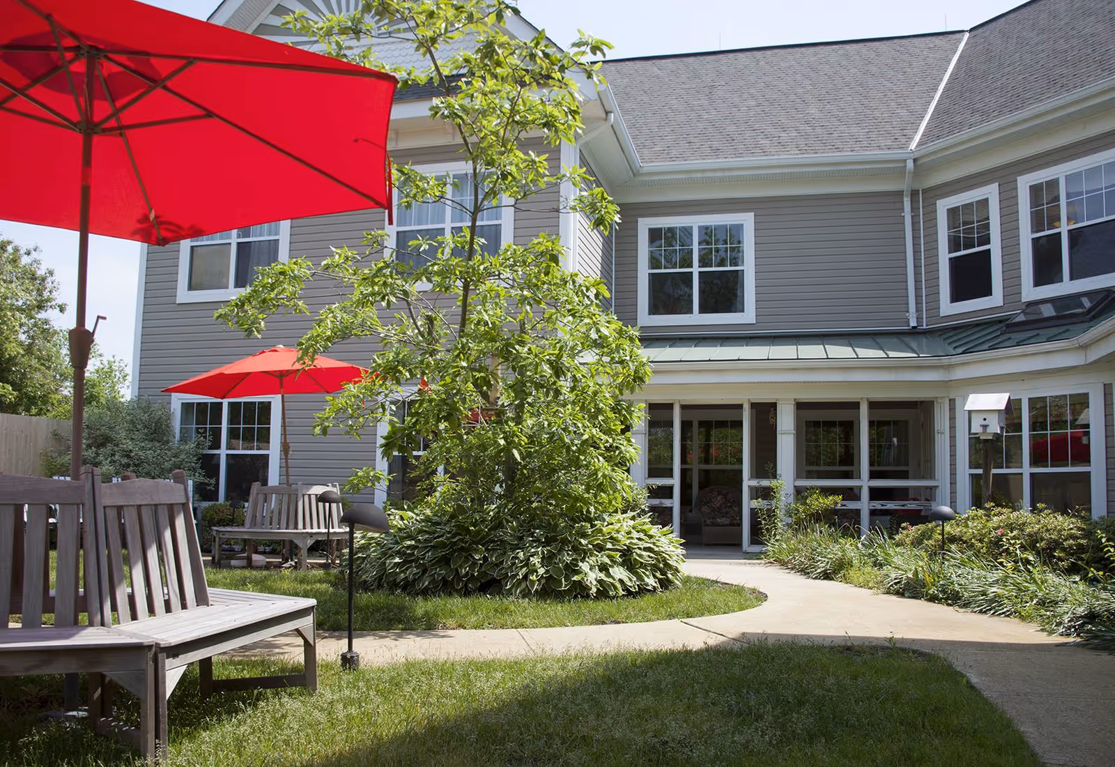 Outdoor courtyard area of a senior living facility with wooden benches, red umbrellas, a tree surrounded by plants, and a two-story building with multiple windows in the background.
