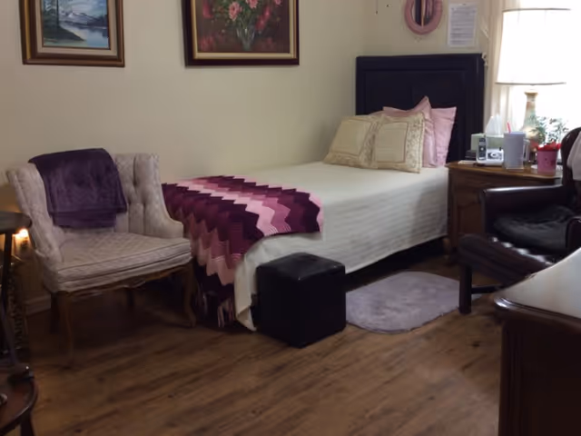 A cozy bedroom with a single bed covered in a white blanket and a purple and pink crocheted throw. There are two decorative pillows on the bed and a black cushioned headboard. Next to the bed is a wooden nightstand with a lamp, a phone, and some small items. To the left of the bed is a beige upholstered armchair with a purple throw draped over it. The room has wooden flooring and two framed paintings hanging on the wall above the bed and chair.