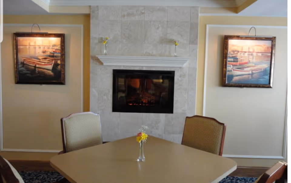 A cozy interior room featuring a beige table with a small vase of yellow and white flowers in the center, surrounded by cushioned chairs. The background shows a tiled fireplace with a white mantel, flanked by two framed paintings of boats on water, each with a small yellow flower in a vase on the mantel above.