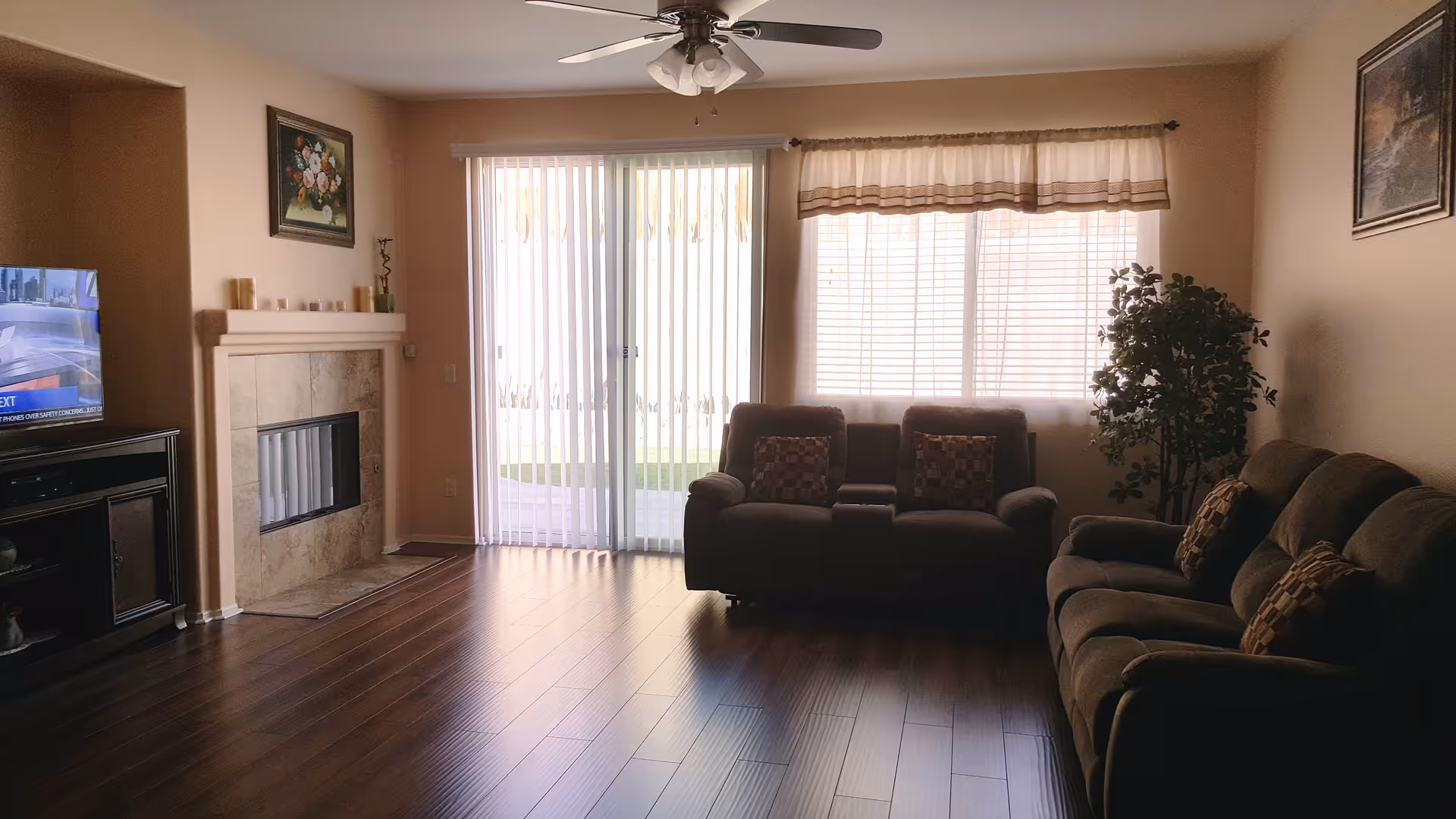 A living room with wooden flooring, a ceiling fan with lights, a fireplace with a painting above it, a television on a stand, two brown sofas with patterned cushions, a large window with blinds and a valance, a sliding glass door with vertical blinds, and a potted plant in the corner.