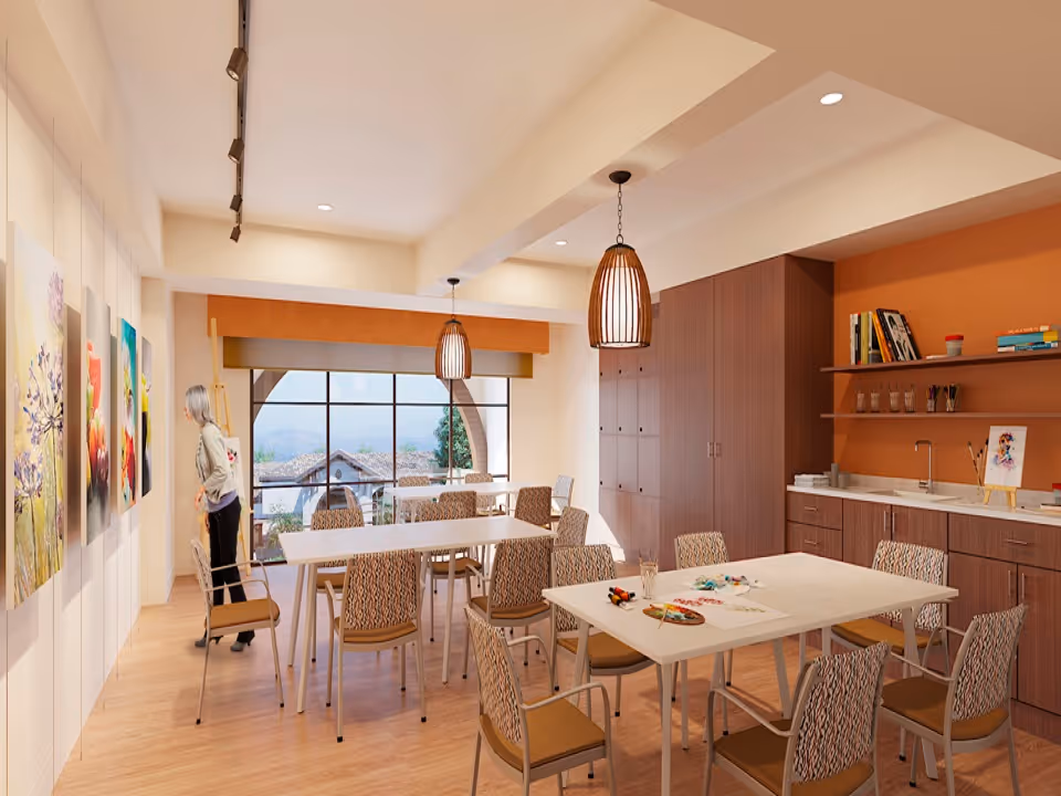 A bright and spacious art room with several tables and chairs arranged for group activities. A woman is observing colorful paintings displayed on the wall. The room features large windows with a scenic view outside, wooden cabinetry, a countertop with art supplies, and warm pendant lighting.