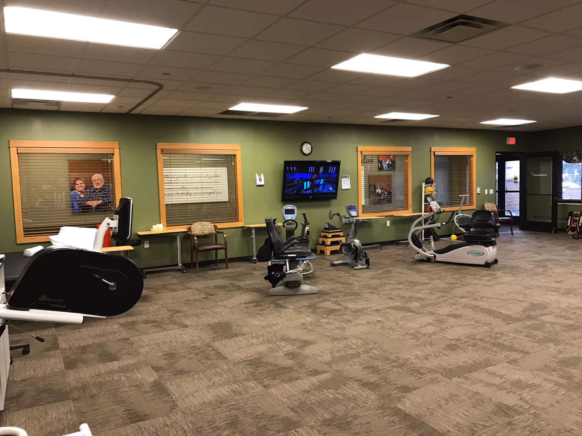A spacious exercise room in a senior living facility with several stationary exercise bikes and fitness equipment. The room has green walls with wooden framed windows, a wall-mounted TV, a clock, and chairs along the walls. There is a door leading outside or to another area of the facility.