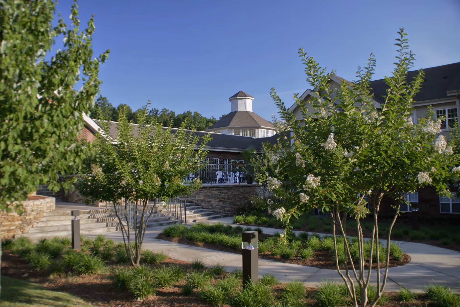 Outdoor view of The Haven at Regency Pointe showing a landscaped garden with small trees and shrubs, paved walkways, and a building with a stone facade and a cupola under a clear blue sky.