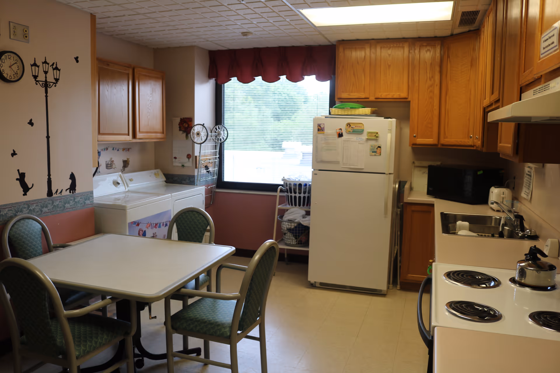 A kitchen and laundry area in a senior living facility with a white refrigerator, wooden cabinets, a stove with a kettle, a microwave, a sink, and a table with four green cushioned chairs. There is a washing machine and dryer against the wall, a window with blinds and a red valance, and wall decorations including a clock and a street lamp decal with cats and butterflies.