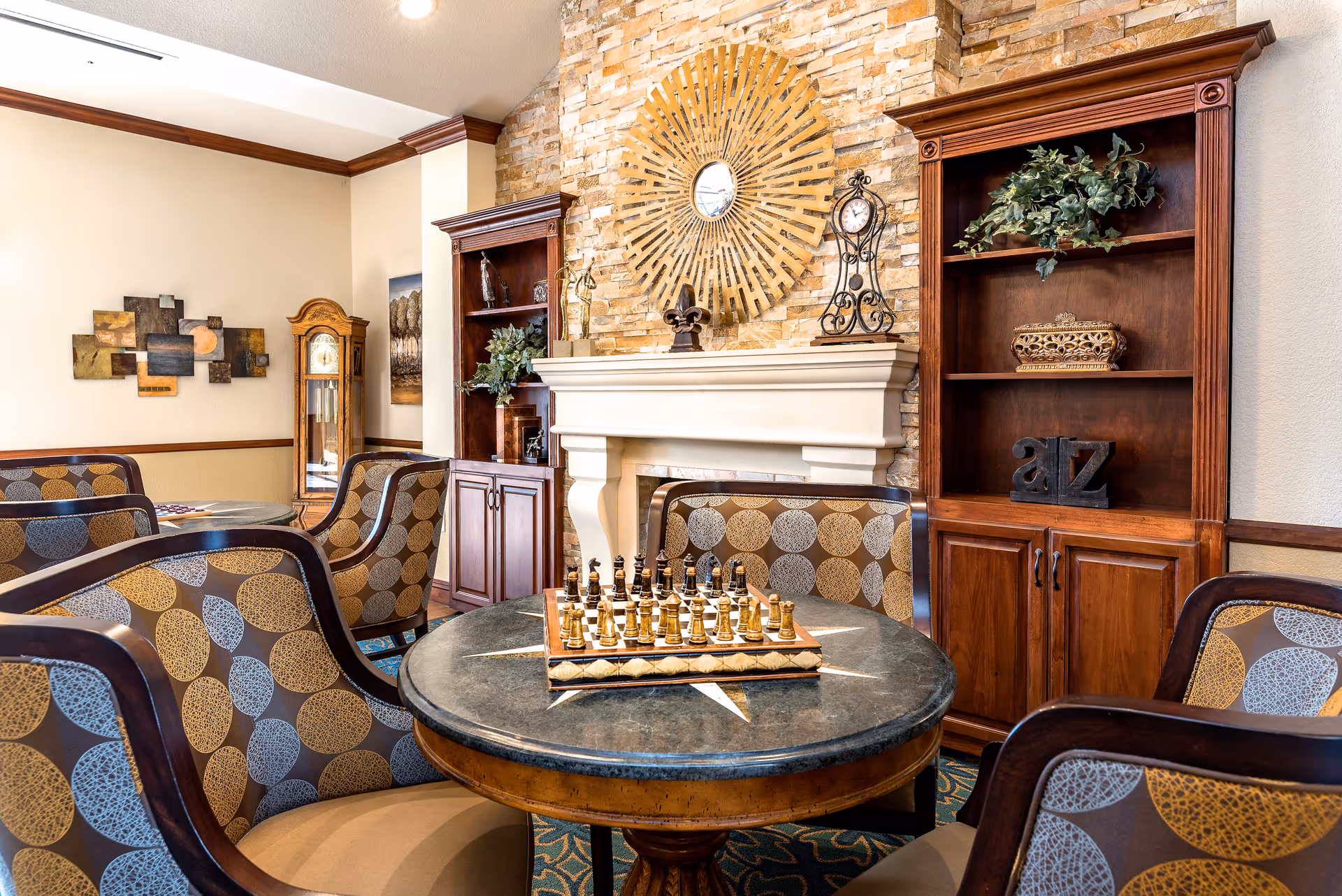 Cozy common room with a round table and chess set, upholstered chairs, a fireplace and decorative shelving.