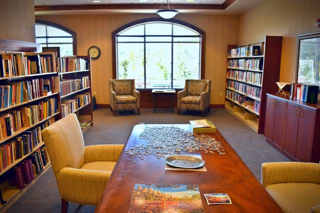 A cozy library room with bookshelves filled with books on both sides. In the center, a wooden table holds a partially completed jigsaw puzzle and some puzzle boxes. Two comfortable armchairs are positioned near a large window with an arched top, letting in natural light. A clock is mounted on the wall to the left.