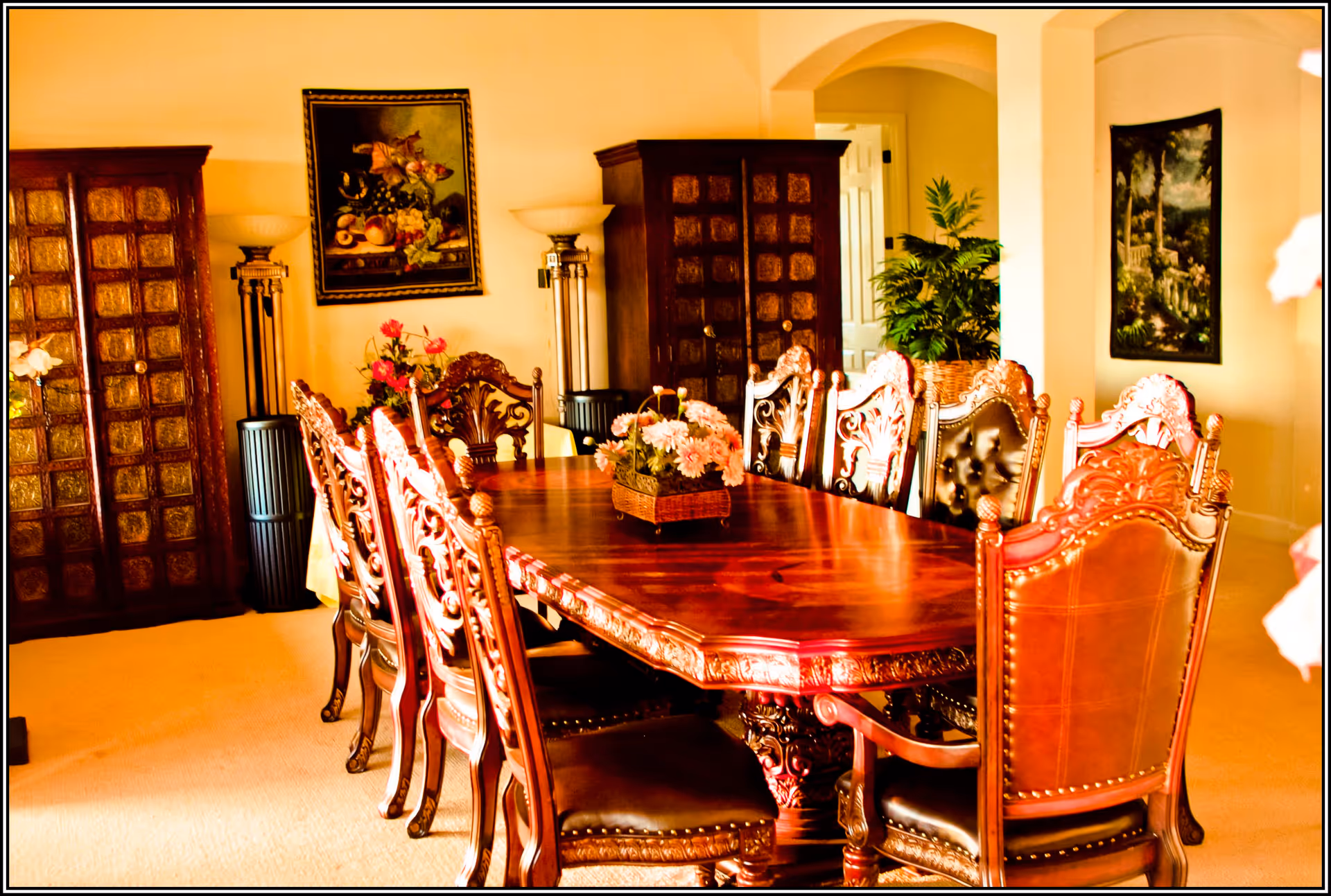 Ornate wooden dining table with carved chairs and a floral centerpiece in a warmly lit dining room.