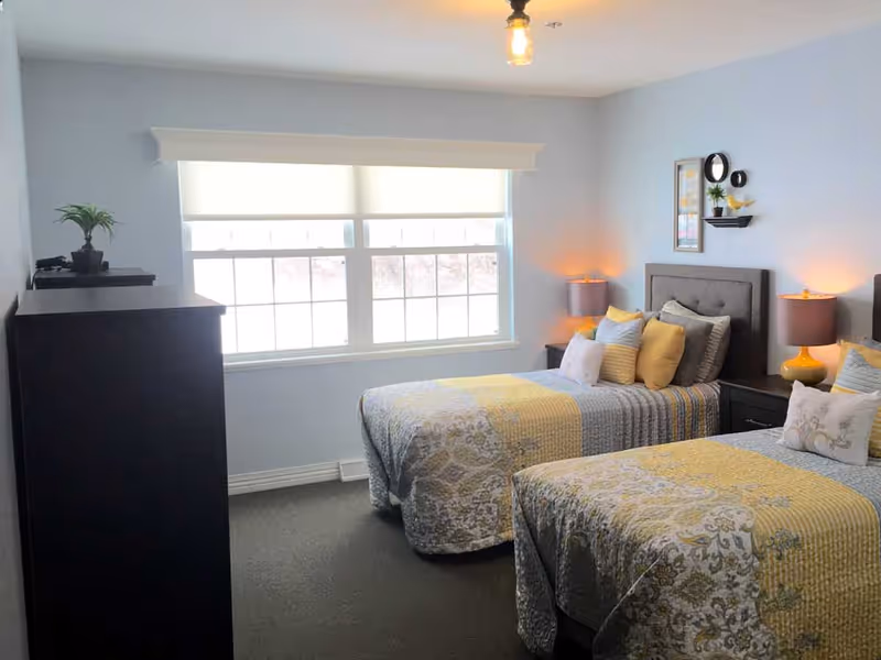 A bright bedroom with two twin beds, each with patterned yellow and gray bedding and multiple pillows. Between the beds is a nightstand with a yellow lamp on each side. A large window with a white blind lets in natural light. There is a dark dresser with a small plant on top against the left wall. The walls are painted light blue and decorated with framed art and small shelves.