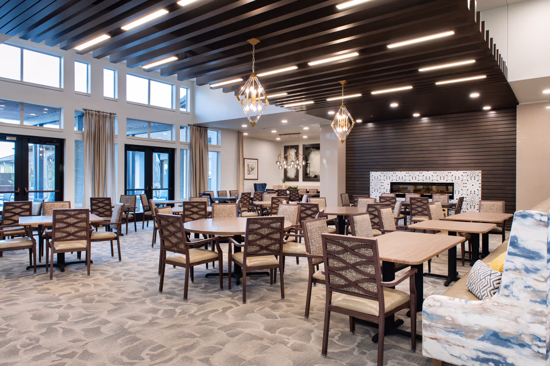 A spacious and modern dining room in a senior living facility with multiple wooden tables and chairs arranged neatly. The room features large windows with beige curtains, a patterned carpet, decorative pendant lights hanging from a ceiling with dark wooden beams, and a fireplace with a white decorative surround on a dark wood-paneled wall.