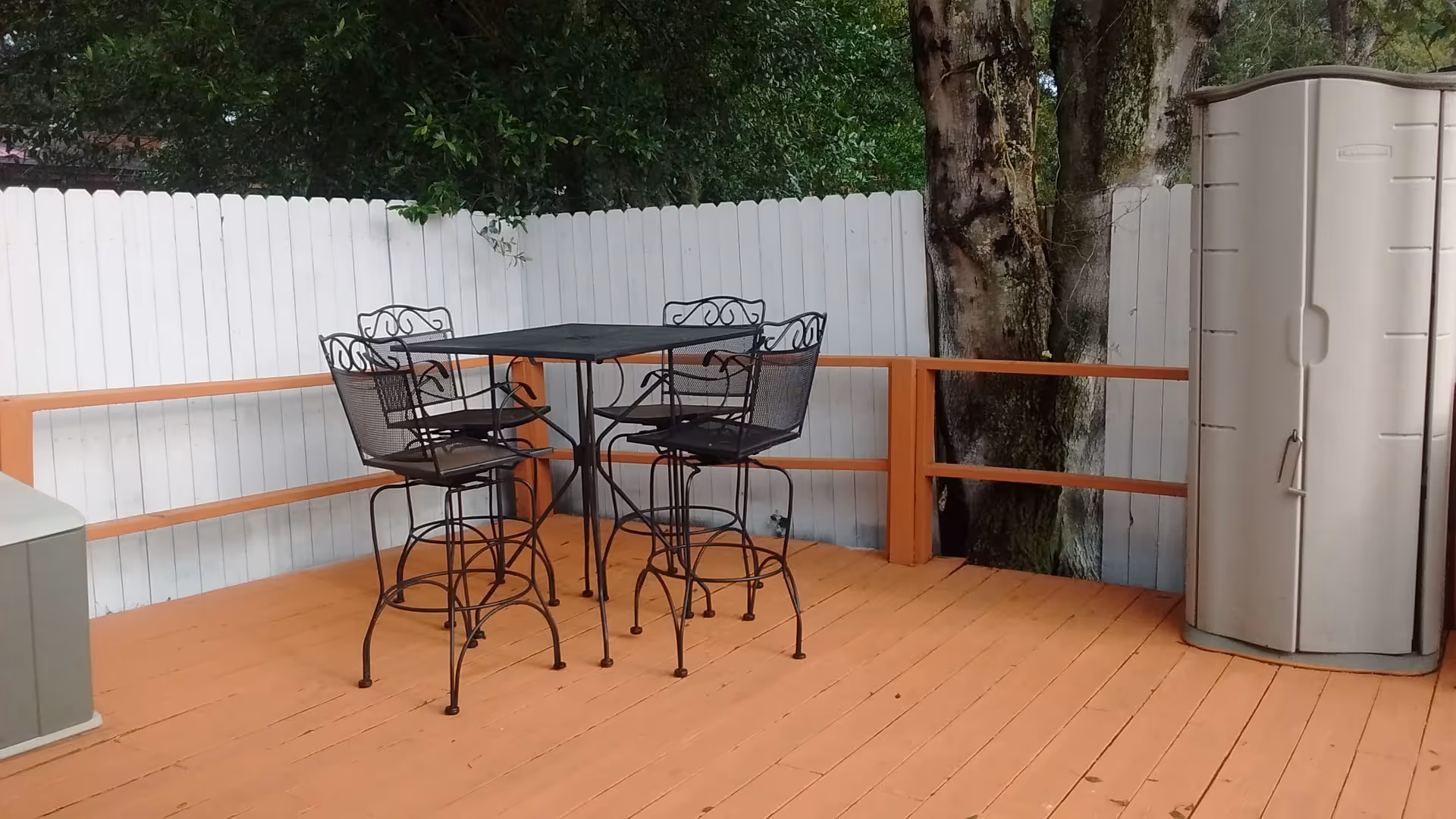 Outdoor wooden deck area with a black metal table and four matching high chairs. The deck is surrounded by a white wooden fence and has trees visible behind it. There is a tall, beige storage cabinet on the right side of the deck.