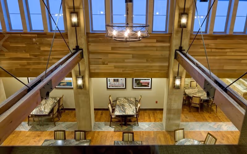 Interior view of a dining area with multiple tables and chairs arranged neatly. The room features wooden floors, large wooden beams, and a high ceiling with skylights. Wall-mounted lights and framed pictures decorate the walls.