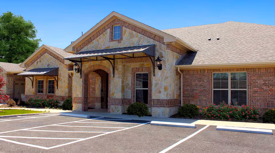 Front exterior of a single-story stone-and-brick memory care building with an entrance awning, windows, landscaping, and parking spaces.