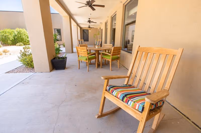 Covered outdoor patio at a senior living facility featuring a wooden rocking chair with a striped cushion and a round table with chairs under ceiling fans.