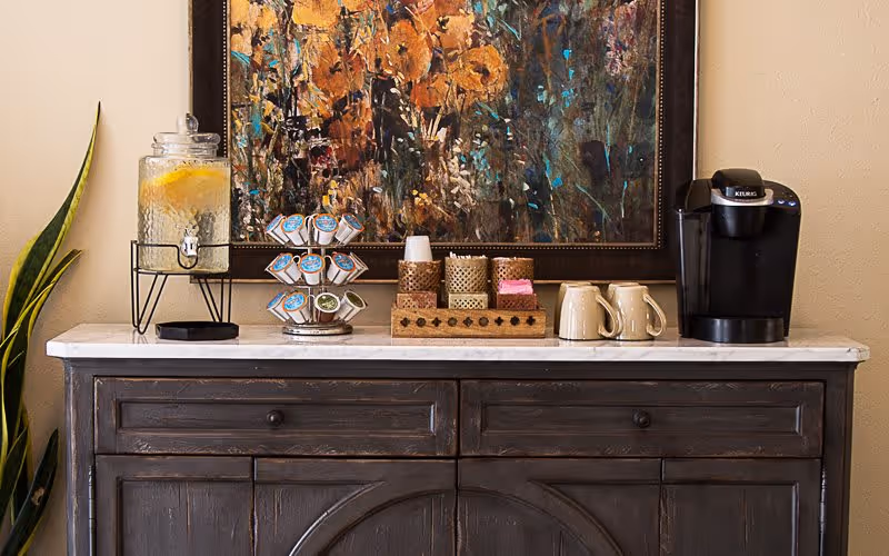 A coffee and beverage station on a wooden sideboard with a Keurig, mugs, K-Cups, and a lemon water dispenser beneath a framed painting.