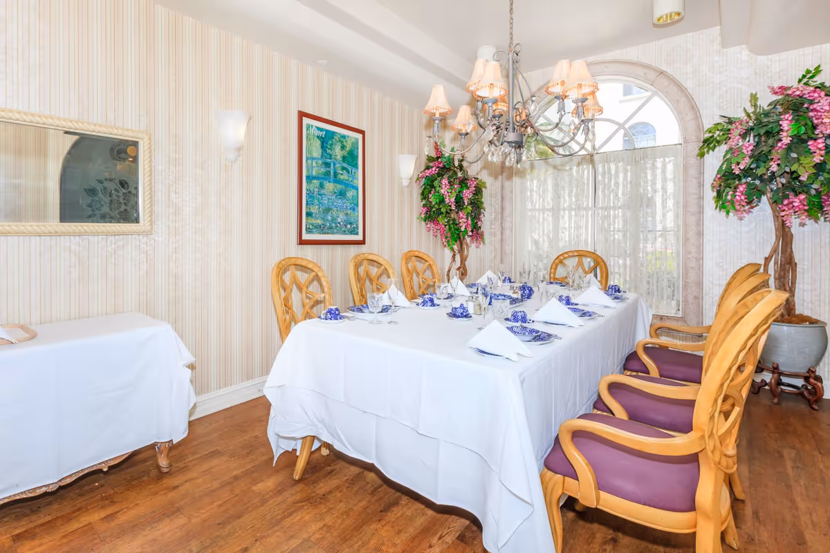 A formal dining room with a long table covered with a white tablecloth, set with blue and white china, glassware, and folded white napkins. The room features wooden chairs with purple cushions, a chandelier with lampshades hanging above the table, floral arrangements in large pots, a framed painting on the wall, and a window with sheer curtains allowing natural light in.