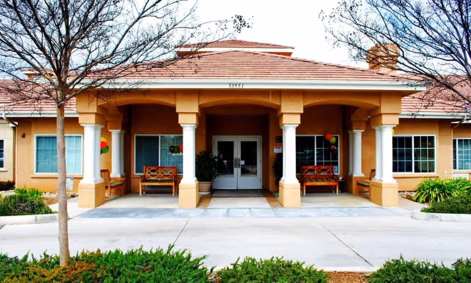 Front exterior view of Wildwood Canyon Villa Assisted Living and Memory Care building with a covered entrance supported by columns, benches on either side, and some trees and shrubs in the foreground.